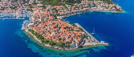 An aerial view of a coastal town with a prominent square surrounded by buildings with red-tiled roofs, a marina with boats, and a clear blue sea.