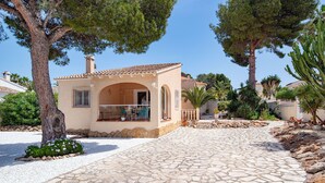 Una villa in stile mediterraneo con un cortile in pietra, un'area salotto esterna coperta e una vegetazione rigogliosa.