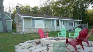 A house with a stone fire pit, four Adirondack chairs, and a grassy yard. - Firefly Cottage | West Twin Lake Getaway (Lewiston)