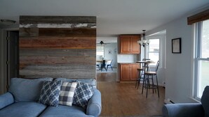 A living room with a blue sofa, wooden wall paneling, and a kitchen in the background. - Firefly Cottage | West Twin Lake Getaway (Lewiston)