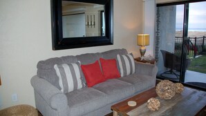 A living room with a grey sofa, red and striped pillows, a wooden coffee table, a mirror, and a view of the beach through a glass door. - On the Promenade 1st floor. Spectacular Panoramic Ocean Views, Patio with grassy (Seaside)