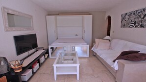 Un espacio habitable pequeño con televisor, un sofá blanco, una mesita de centro y una cama con armario abierto.