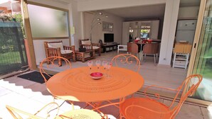 Un comedor con una mesa y sillas naranjas, un televisor al fondo y una cocina visible a través de una puerta abierta.