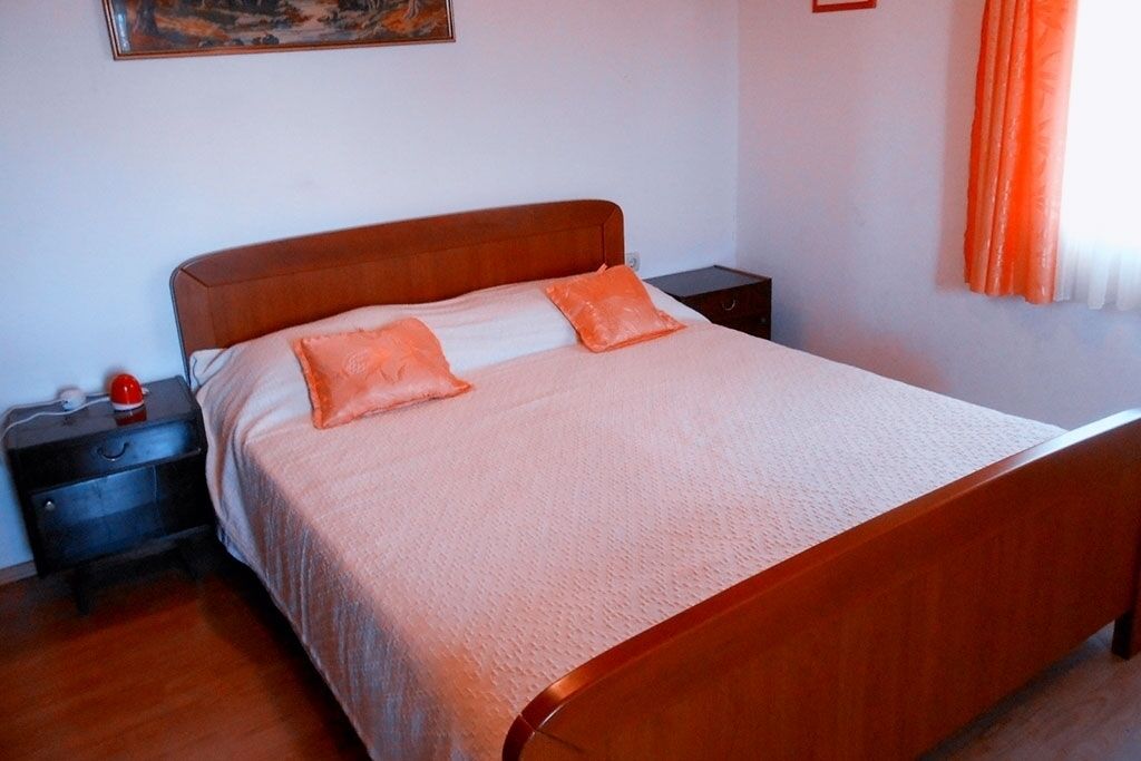 A neatly made bed with a wooden headboard and two pillows, a nightstand with a red object on it, and a painting on the wall.