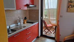 Une cuisine avec des placards rouges, un évier et une fenêtre donnant sur un balcon où se trouve une chaise.