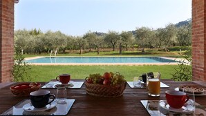A wooden table with breakfast items and a view of a swimming pool and olive trees.