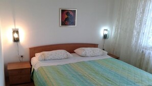 A single bed with a green quilt, two bedside lamps, a nightstand, and a painting on the wall.
