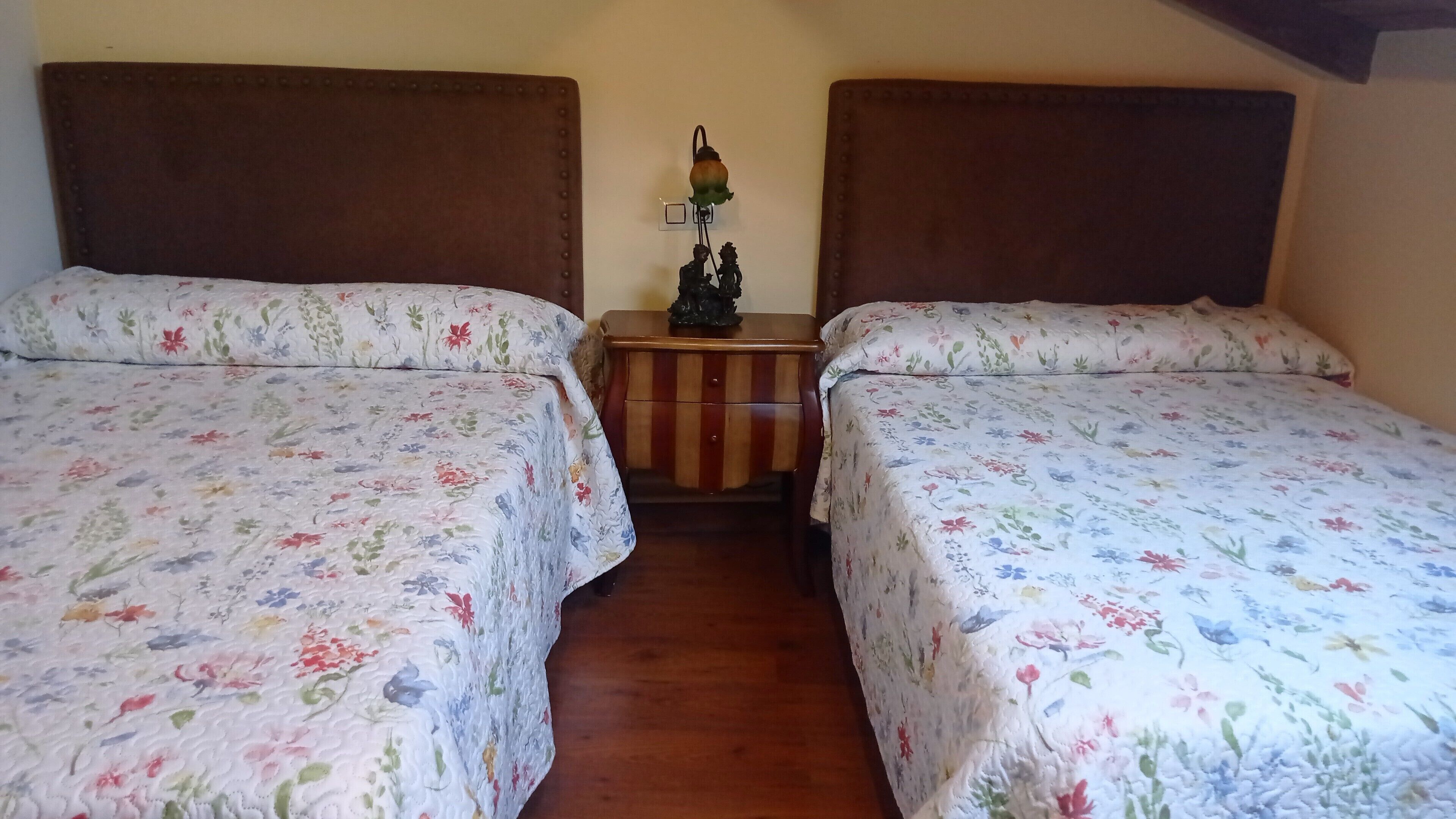 Ein Zimmer mit zwei Betten, jeweils mit blumigem Bettbezug, einem kleinen Nachttisch mit einer Dekoration und einer Wandleuchte.