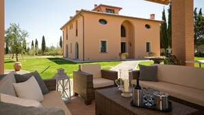 A covered outdoor seating area with wicker furniture and a table set for dining, overlooking a large building with arched windows and a garden. - Villa for 11 People in Tuscany with private pool (Peccioli)
