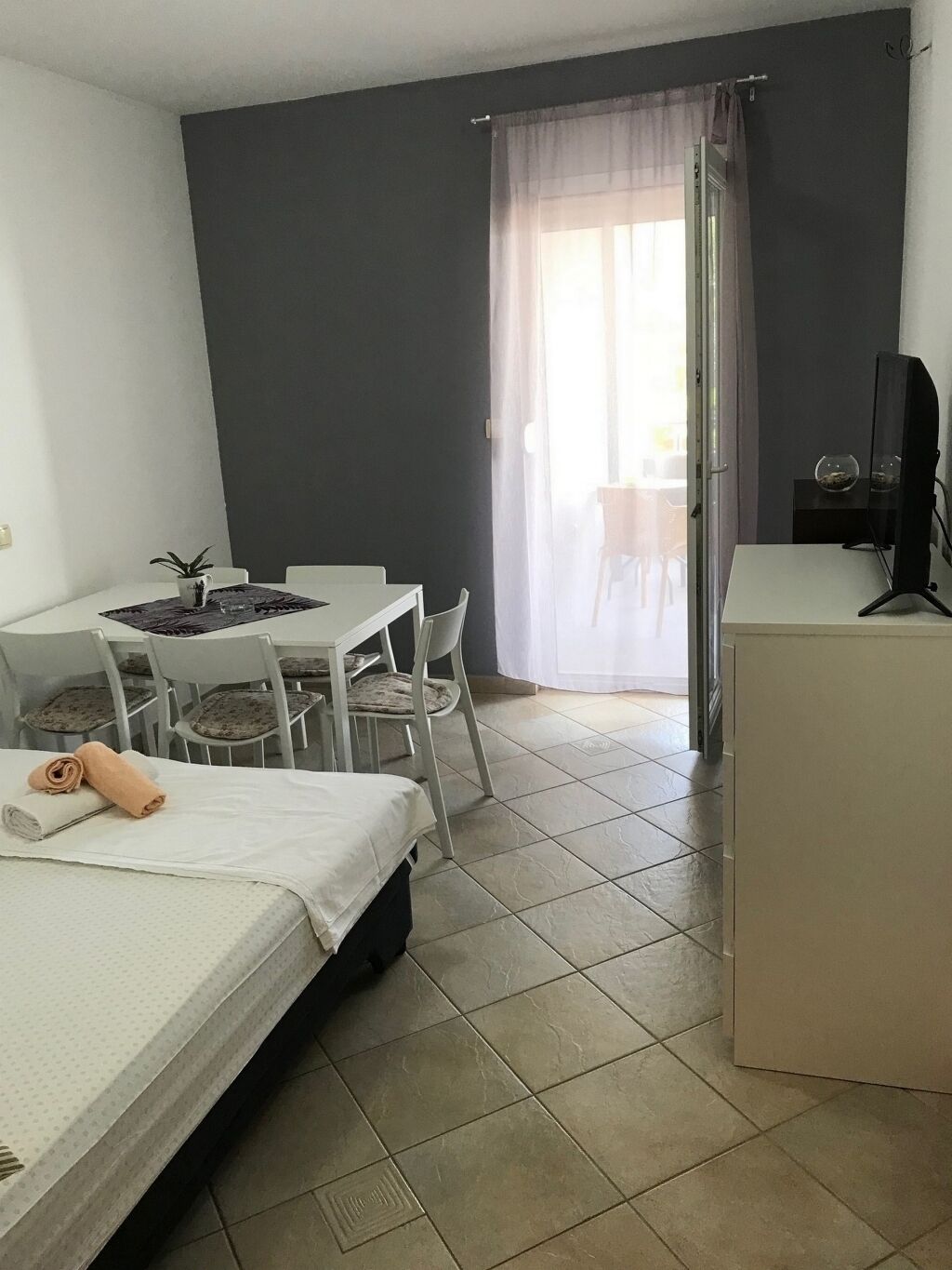 A small, well-lit room with a bed, a desk, a chair, a television, and a window with curtains.