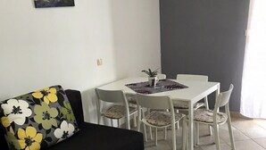 Ein Wohnzimmer mit einer schwarzen Couch, einem weißen Esstisch mit vier Stühlen, einem kleinen Topfpflanzen und einem Bild an der Wand.