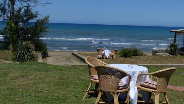 Ein Außenbereich mit Korbstühlen und einem Tisch, mit Blick auf einen Strand, Wellen und einen klaren Himmel.