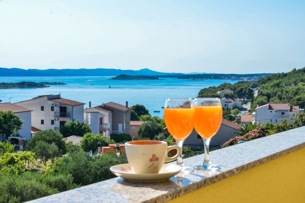 Un balcon, une tasse de café et deux verres de jus d’orange, avec vue sur une ville côtière, ses bâtiments et la mer.