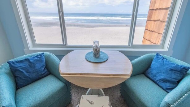 Una habitación con un ventanal grande que da a una playa, dos sillas color turquesa, una mesita de madera con un florero y un muro de listones de madera.