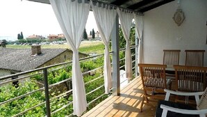 A balcony with white curtains, wooden chairs, and a view of a green landscape.