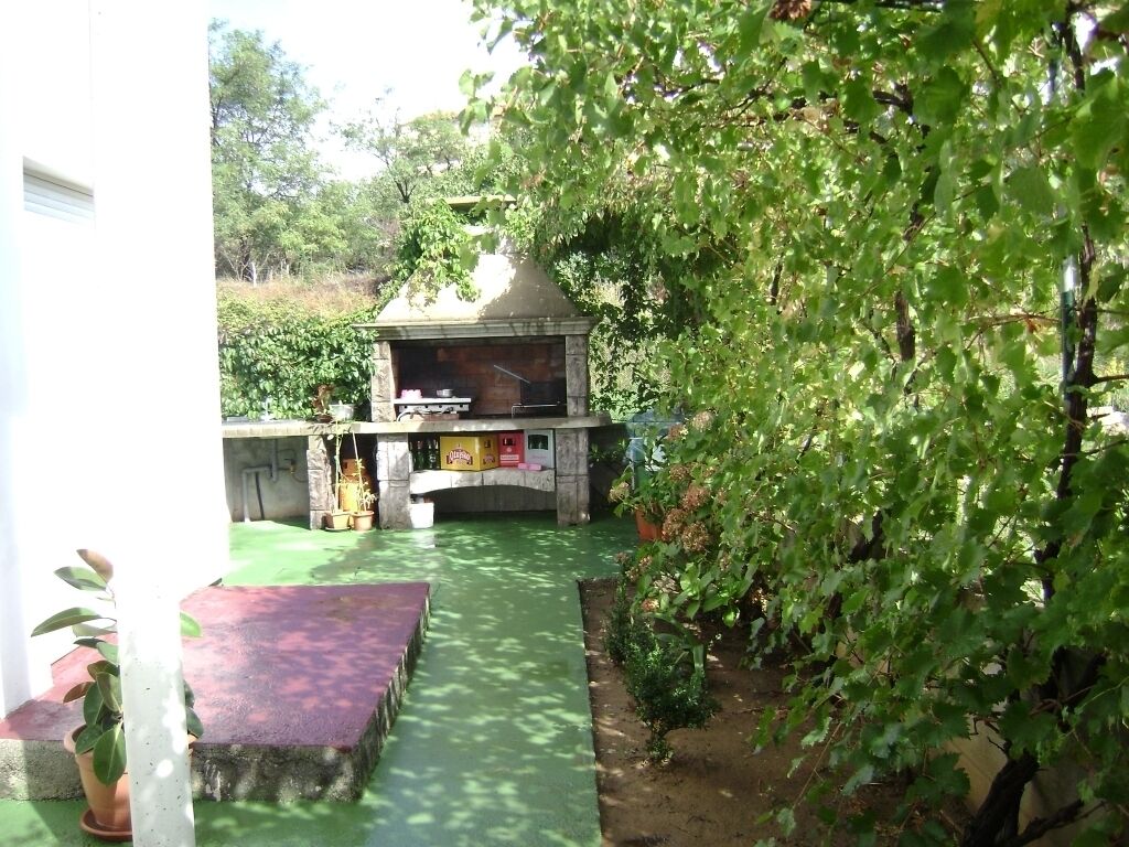 A patio area with a barbecue grill, surrounded by greenery and a small bridge.