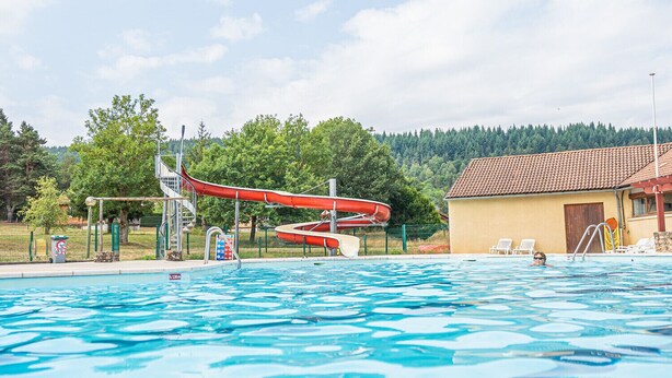 Ein Schwimmbecken mit Wasserrutsche, einem Rettungsturm und einem kleinen Gebäude mit einem Ziegeldach.
