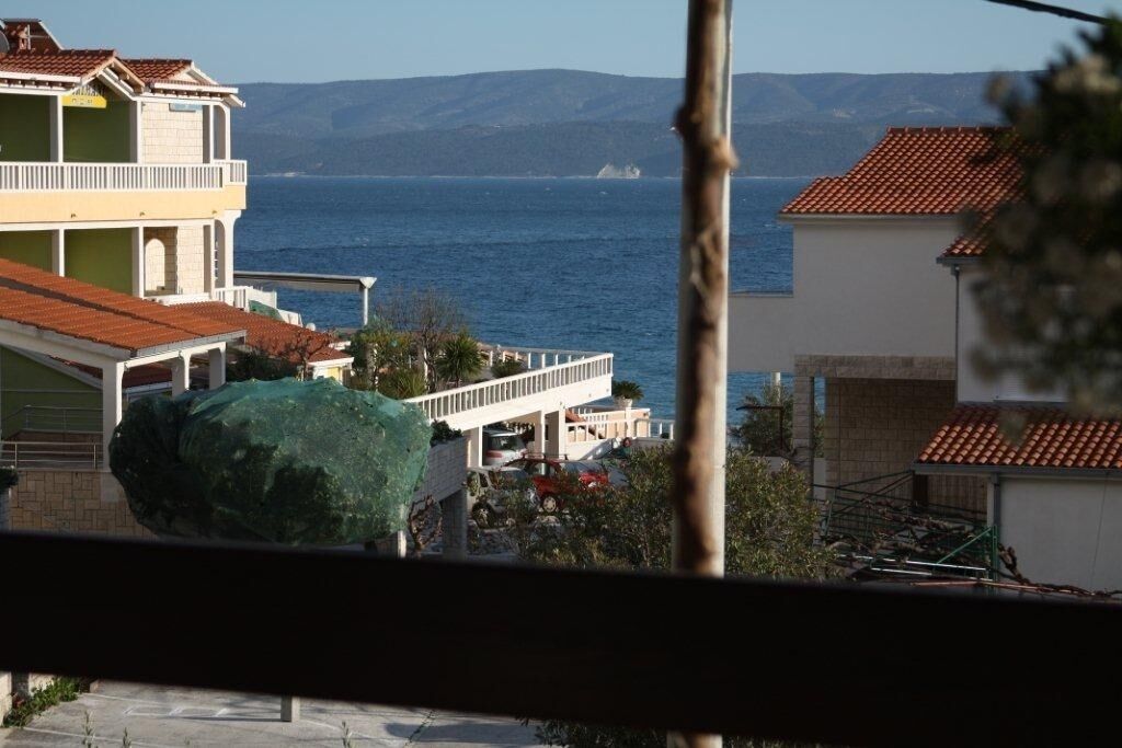 A coastal building with a balcony overlooking the sea, a large rock in the foreground, and a mountainous backdrop.