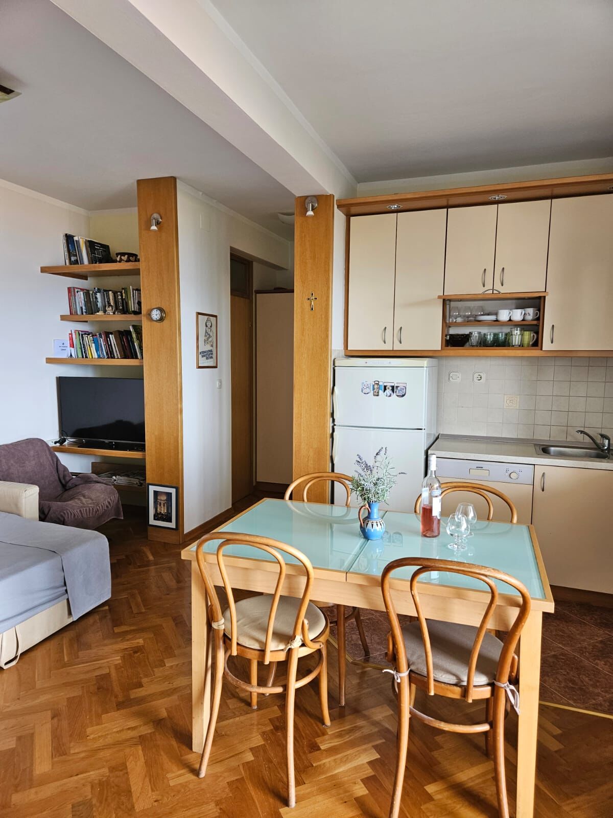 Ein kompakter Wohnbereich mit Esstisch, Stühlen, einer Couch, einem Bücherregal, einem Fernseher und einer Küchenzeile inklusive Schränken und Kühlschrank.