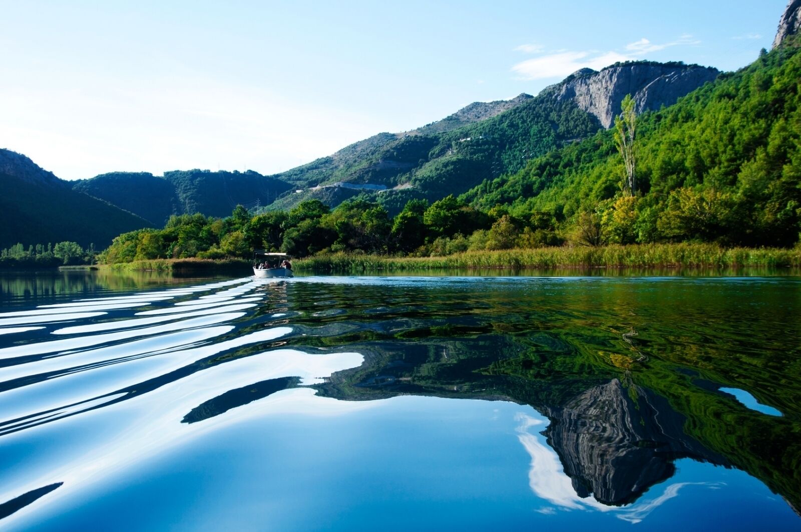 A calm lake with clear reflections, surrounded by lush green hills and a boat in the distance.