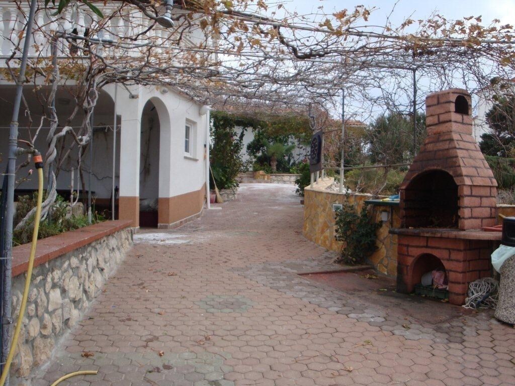 A paved pathway leading to a building with a brick oven, surrounded by vegetation and a stone wall.