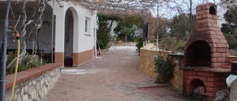 A paved pathway leading to a building with a brick oven, surrounded by vegetation and a stone wall.