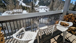 Un balcón con vista a una montaña nevada y un río. Hay dos sillas de plástico blanco y una mesa pequeña con una pila de leña.