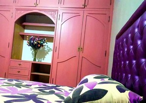 A bedroom with a pink wardrobe, a bed with a floral bedspread, and a purple headboard.