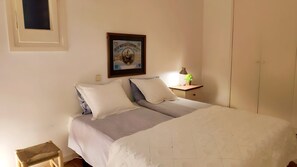 A bedroom with a bed, pillows, a bedside table with a lamp, a framed picture on the wall, and a small stool.