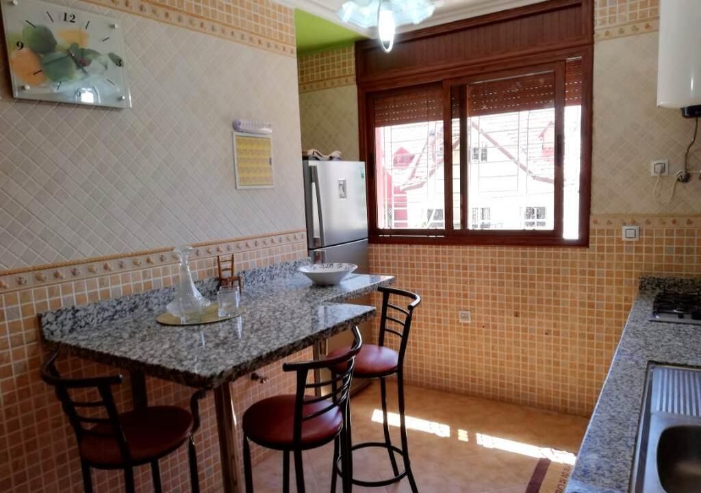 A kitchen with a granite countertop, two bar stools, a refrigerator, and a window with blinds.