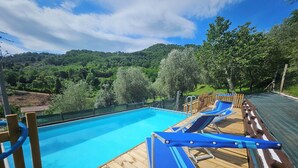 Piscina con muelle de madera y sillones de descanso, rodeada de vegetación y colinas.