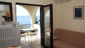 Een woonkamer met een bank, een salontafel en een televisie. Er is een balkon met een tafel en stoelen, en uitzicht op de zee.