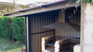 Zona de barbacoa a l'aire lliure d'estil rústic, amb xemeneia, envoltada per una estructura de formigó i un teulat metàl·lic.