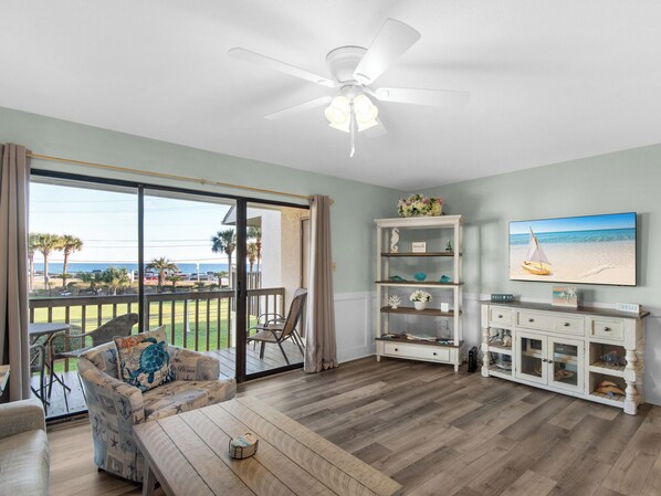 A spacious living room with a large sliding glass door leading to a balcony, a ceiling fan, a television mounted on the wall, and a wooden coffee table.