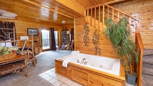Interior de uma cabana de madeira com uma grande banheira de madeira, uma área de refeições com mesa e cadeiras, e uma escada que leva a um andar superior.