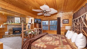 Um quarto de cabana de troncos aconchegante com lareira, uma cama grande com colcha, um ventilador de teto e uma parede de pedra.