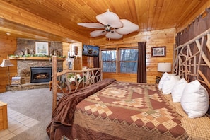 Ein gemütliches Schlafzimmer in einer Blockhütte mit Kamin, einem großen Bett mit Patchworkdecke, einem Deckenventilator und einer Steinwand.