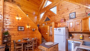 Interior acolhedor de uma cabana, com paredes de madeira, área de jantar, cozinha com frigorífico e máquina de lavar loiça, e janela que proporciona luz natural.