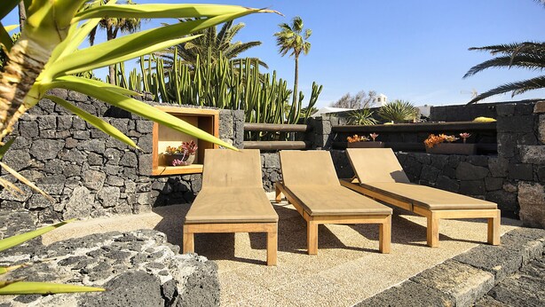 Un espacio al aire libre iluminado por el sol con sillones reclinables y un muro de piedra como fondo, con cactus y una chimenea de piedra.