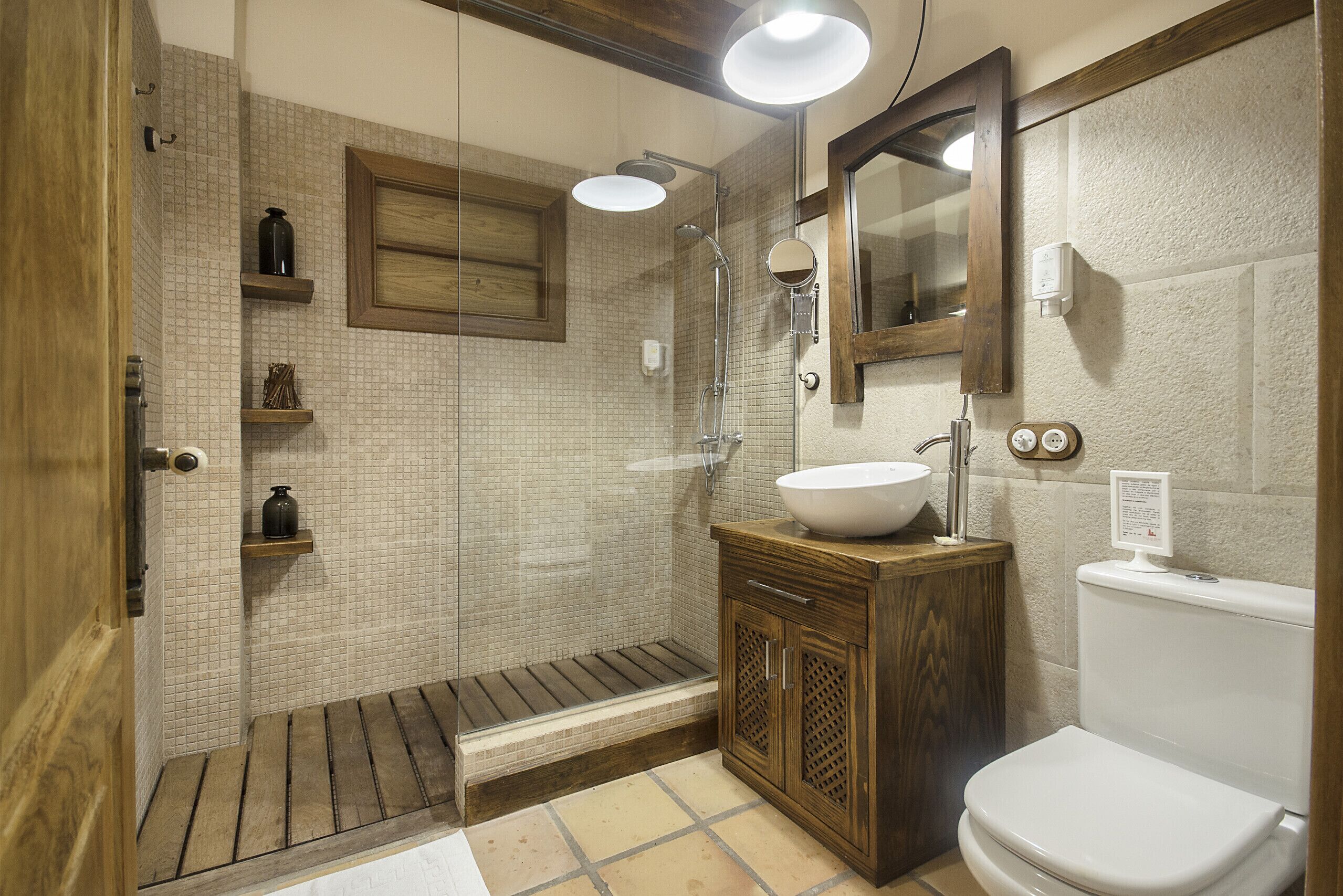 Un baño con tocador de madera, un lavabo blanco, un inodoro, una ducha con puerta de vidrio y un espejo sobre el lavabo.