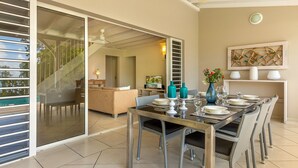 Comedor con mesa de vidrio, sillas de mimbre y vistas a una piscina y zona de estar exterior.