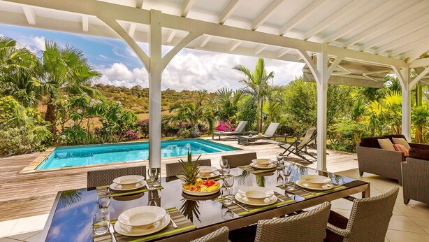 Un área de comedor al aire libre techada con vista a la piscina, mobiliario de mimbre y un entorno de jardín tropical.