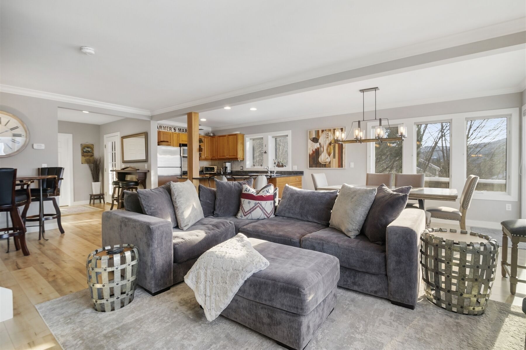 A spacious living room with a large grey sectional sofa, a dining area with wooden chairs, a kitchen with wooden cabinets, and a view of trees through large windows.