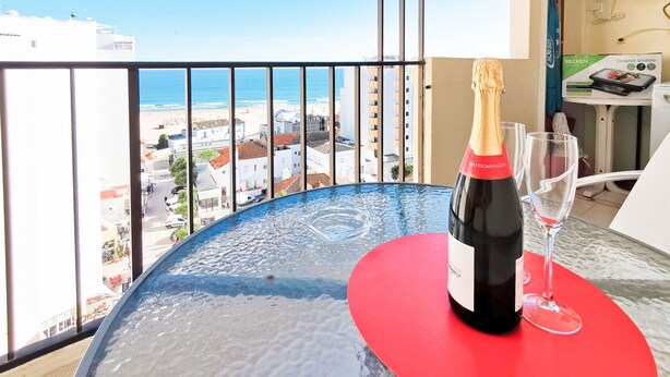 A balcony with a table, a bottle of champagne, and a glass. The view includes a beach and buildings.