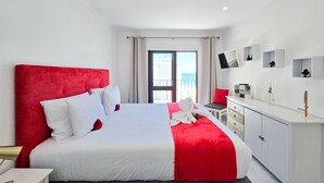 A modern hotel room with a large bed, a desk, and a view of the sea.