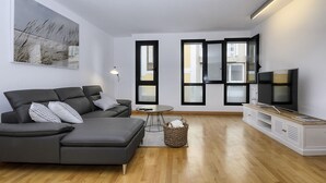 Una sala moderna con un sofá seccional gris, un televisor sobre un mueble de madera y un ventanal grande con marcos negros.
