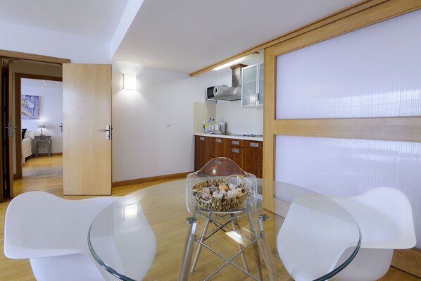 A modern dining area with a glass table and white chairs, a clear plastic bowl on a stand, and a kitchen in the background.