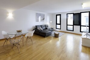 A modern living room with a grey sofa, white chairs, a glass table, and a wooden floor.