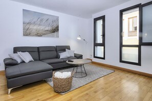 Ein modernes Wohnzimmer mit einem grauen Ecksofa, einem runden Couchtisch und einem großen Fenster mit Jalousien.