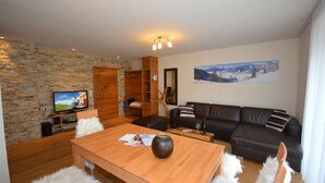 A cozy living room with a stone wall, a wooden entertainment unit, a leather sofa, and a large window with a mountain view. - Zell City Exclusive Lodges - TOP 4 (Zell am See)